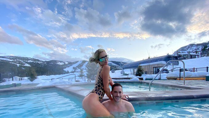 Paris is seen posing with Carter, who sits against a snowy backdrop, to his shoulders at a swimming pool in an animal print swimsuit at yellowstone club